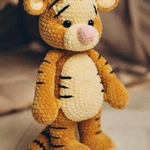 May include: A plush tiger toy made of soft, textured orange and cream yarn. The stuffed animal features black embroidered stripes on its body, arms, legs, and tail, a light pink nose, and small black eyes. It stands upright with a round head.