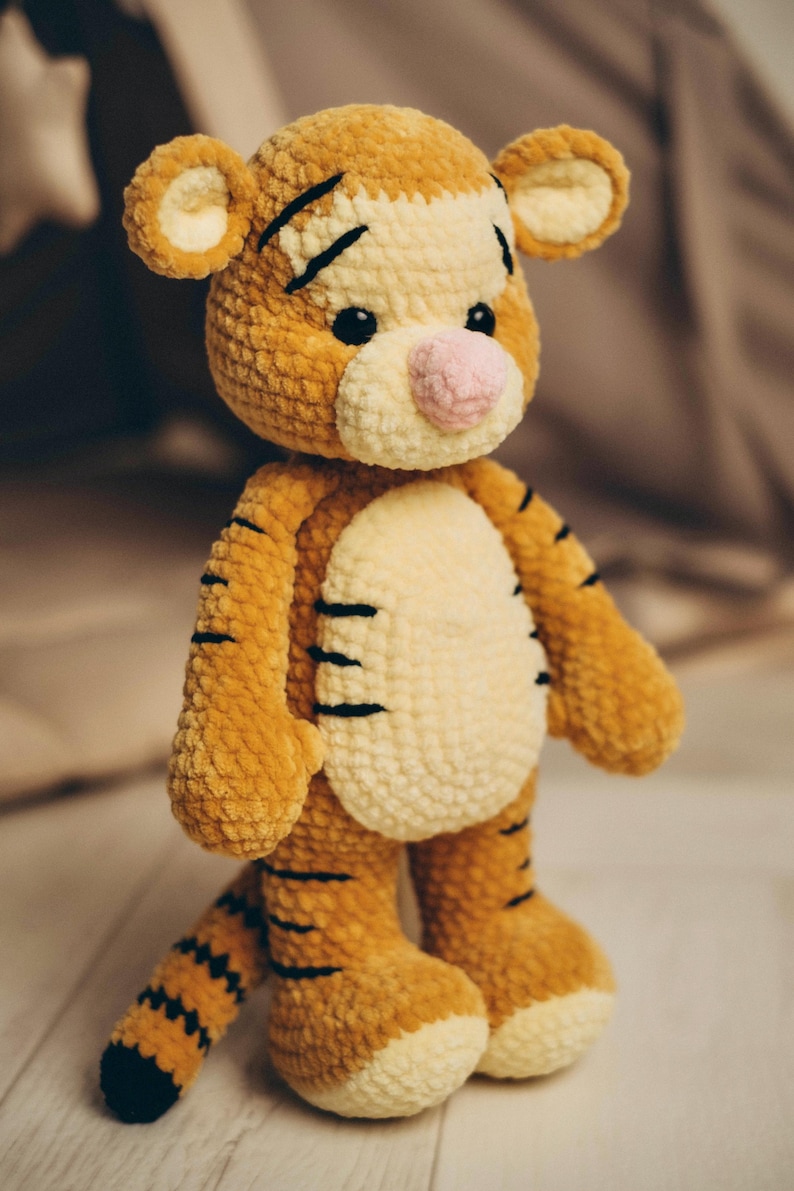 May include: A plush tiger toy made of soft, textured orange and cream yarn. The stuffed animal features black embroidered stripes on its body, arms, legs, and tail, a light pink nose, and small black eyes. It stands upright with a round head.