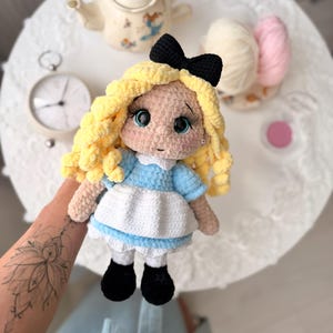 Può includere: Bambola all'uncinetto fatta a mano di Alice nel Paese delle Meraviglie, con un vestito blu con un grembiule bianco, scarpe nere e un fiocco nero. La bambola ha capelli ricci gialli e grandi occhi. Un oggetto artigianale stravagante.