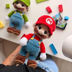 May include: Two handmade crochet plush dolls representing Mario and Luigi. Mario wears a red cap with an M, red shirt, blue overalls with yellow buttons, white gloves, and brown shoes. Luigi wears a green cap with an L, green shirt, and blue overalls.