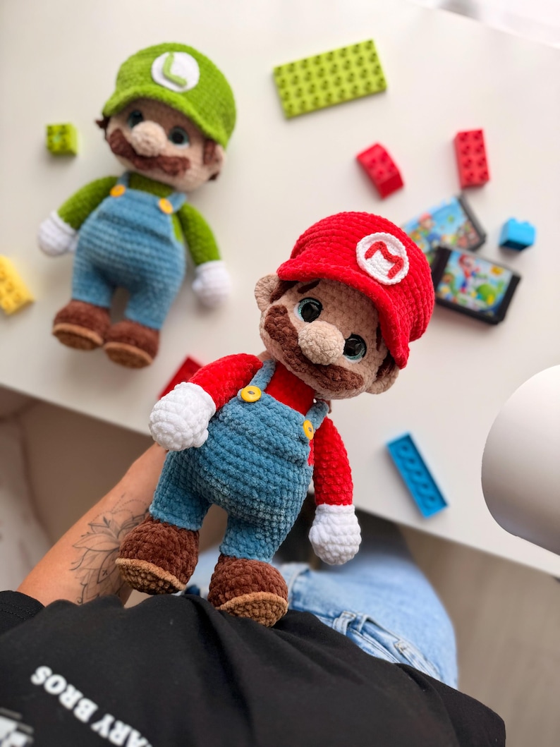 May include: Two handmade crochet plush dolls representing Mario and Luigi. Mario wears a red cap with an M, red shirt, blue overalls with yellow buttons, white gloves, and brown shoes. Luigi wears a green cap with an L, green shirt, and blue overalls.