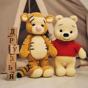 May include: Two crocheted plush toys, a yellow and orange striped tiger and a light yellow bear wearing a red shirt, standing next to a vertical stack of six wooden blocks featuring the Cyrillic letters Д, Р, У, З, Ь, Я.