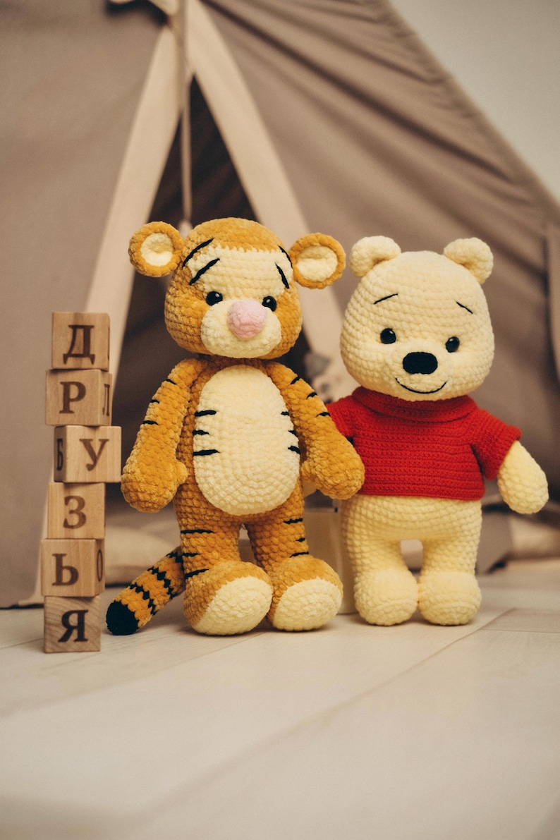 May include: Two crocheted plush toys, a yellow and orange striped tiger and a light yellow bear wearing a red shirt, standing next to a vertical stack of six wooden blocks featuring the Cyrillic letters Д, Р, У, З, Ь, Я.