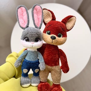 May include: Two handmade crochet stuffed animals: a gray bunny with pink-lined ears and a red fox. The bunny wears a blue and gray outfit, while the fox has tan legs. Both have large, expressive eyes and are standing together.