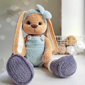 May include: A crocheted plush rabbit with tan fur, long ears, and dark eyes. It wears a light blue headband with a bow, a light blue overall with two small buttons, and dark gray boots. The text PARKAEVA_HM is visible on the right side.