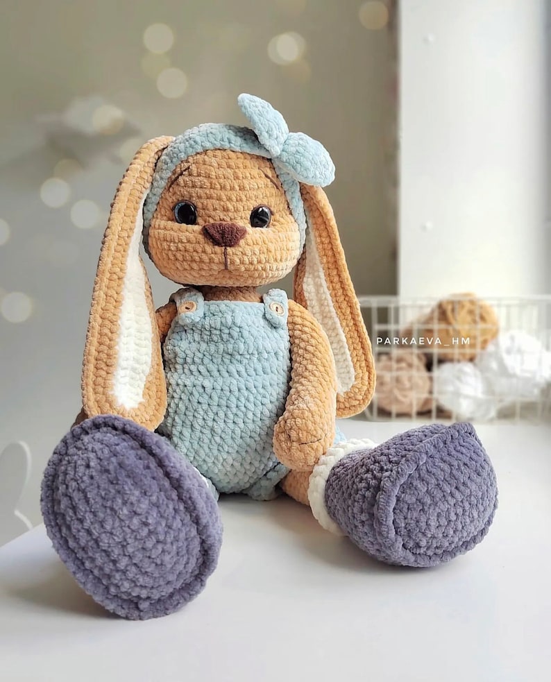 May include: A crocheted plush rabbit with tan fur, long ears, and dark eyes. It wears a light blue headband with a bow, a light blue overall with two small buttons, and dark gray boots. The text PARKAEVA_HM is visible on the right side.