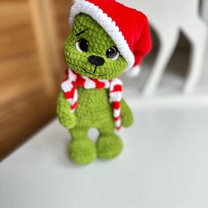 Peut inclure: Une poup&eacute;e crochet&eacute;e &agrave; la main avec un corps textur&eacute; vert, de grands yeux sombres et un nez brod&eacute; noir. La poup&eacute;e porte un bonnet de P&egrave;re No&euml;l rouge vif avec un bord blanc et une &eacute;charpe ray&eacute;e rouge et blanche assortie autour du cou.
