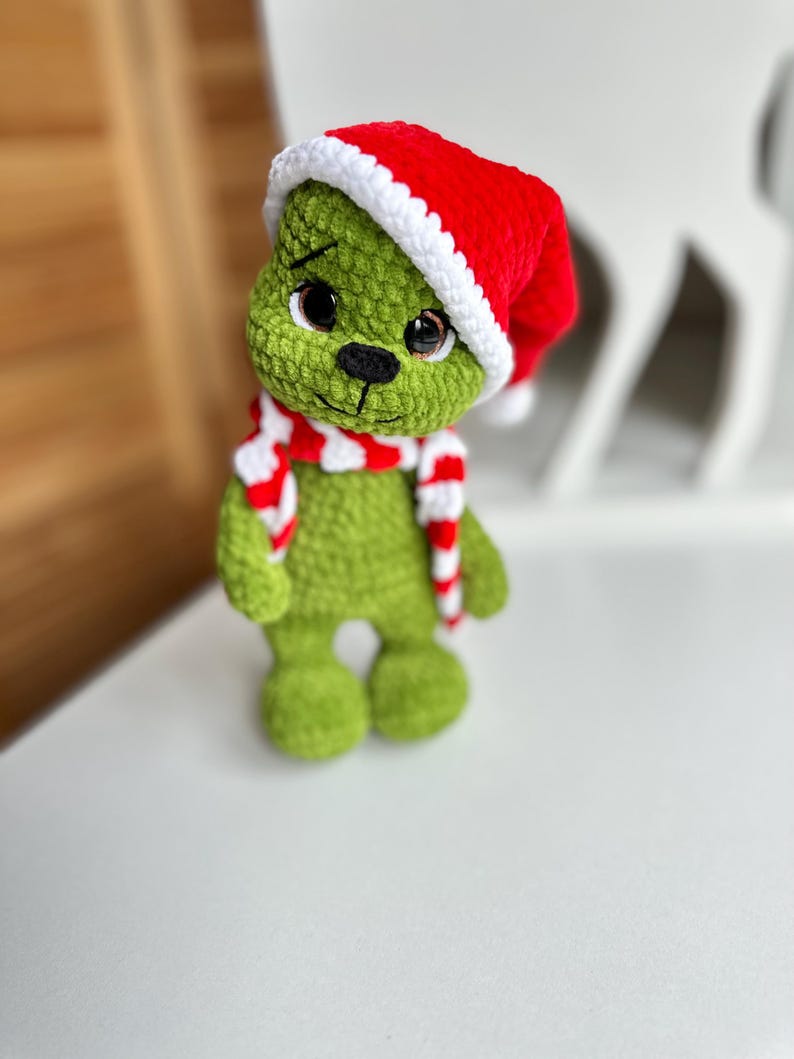 Peut inclure: Une poup&eacute;e crochet&eacute;e &agrave; la main avec un corps textur&eacute; vert, de grands yeux sombres et un nez brod&eacute; noir. La poup&eacute;e porte un bonnet de P&egrave;re No&euml;l rouge vif avec un bord blanc et une &eacute;charpe ray&eacute;e rouge et blanche assortie autour du cou.