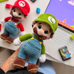 May include: Two handmade crochet plush dolls representing Mario and Luigi. Mario wears a red hat with an M, red shirt, and blue overalls. Luigi wears a green hat with an L, green shirt, and blue overalls. Both have brown mustaches and white gloves.