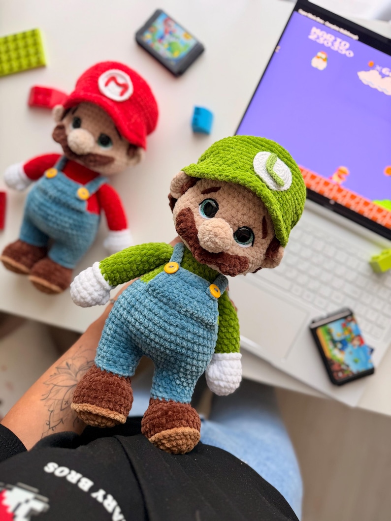 May include: Two handmade crochet plush dolls representing Mario and Luigi. Mario wears a red hat with an M, red shirt, and blue overalls. Luigi wears a green hat with an L, green shirt, and blue overalls. Both have brown mustaches and white gloves.
