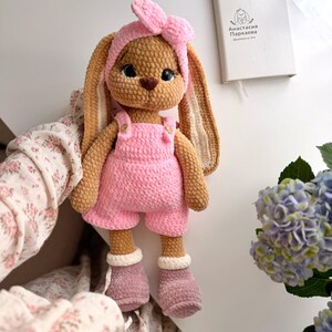 May include: A crocheted stuffed rabbit toy with long ears, wearing a pink headband with a bow, pink overalls with small buttons, and mauve boots with white cuffs. The rabbit has large dark eyes and a brown nose, crafted from textured yarn.