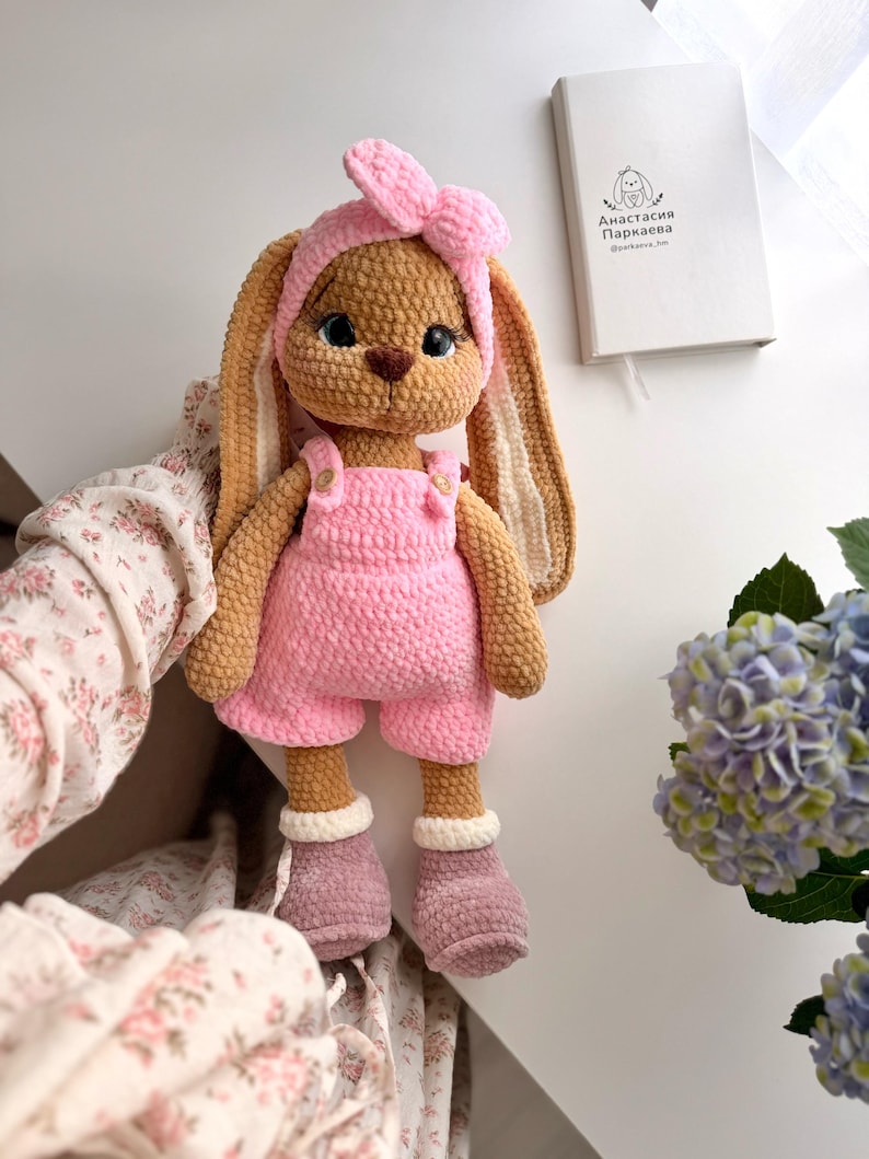 May include: A crocheted stuffed rabbit toy with long ears, wearing a pink headband with a bow, pink overalls with small buttons, and mauve boots with white cuffs. The rabbit has large dark eyes and a brown nose, crafted from textured yarn.