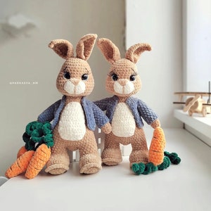 Puede incluir: Dos conejos de crochet que llevan chaquetas azules y sostienen zanahorias. Los conejos son marrones y blancos y tienen orejas largas.