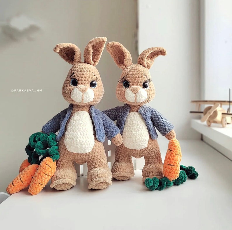 Puede incluir: Dos conejos de crochet que llevan chaquetas azules y sostienen zanahorias. Los conejos son marrones y blancos y tienen orejas largas.
