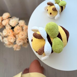 K&ouml;nnte beinhalten: Zwei handgefertigte H&auml;kelbienen-Pl&uuml;schtiere. Die Bienen haben gelbe K&ouml;rper mit braunen und gelben Streifen, wei&szlig;e Fl&uuml;gel und gr&uuml;ne, blattf&ouml;rmige Akzente. Eine Biene befindet sich im Vordergrund, die andere im Hintergrund auf einer wei&szlig;en Oberfl&auml;che.