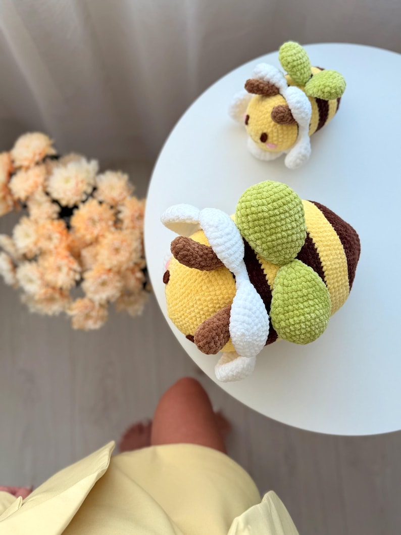 K&ouml;nnte beinhalten: Zwei handgefertigte H&auml;kelbienen-Pl&uuml;schtiere. Die Bienen haben gelbe K&ouml;rper mit braunen und gelben Streifen, wei&szlig;e Fl&uuml;gel und gr&uuml;ne, blattf&ouml;rmige Akzente. Eine Biene befindet sich im Vordergrund, die andere im Hintergrund auf einer wei&szlig;en Oberfl&auml;che.
