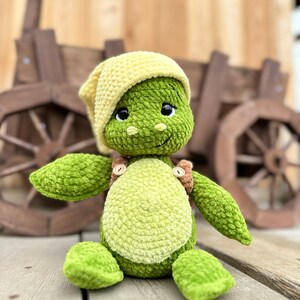 Puede incluir: Un juguete de peluche con forma de tortuga hecho de hilo texturizado verde con vientre verde claro, que lleva un gorro de punto amarillo p&aacute;lido y un peto marr&oacute;n con peque&ntilde;os botones de madera. El peluche tiene grandes ojos negros y boca bordada.