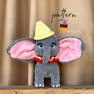 May include: A gray crocheted elephant toy with large pink-lined ears, a yellow hat, and a red ruffled collar. The elephant has blue eyes and is standing on a wooden surface. The word "pattern" is written above the elephant.