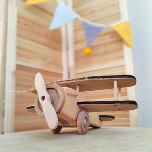 May include: A wooden toy airplane with a propeller, black accents, and wheels. The airplane is sitting on a wooden surface.