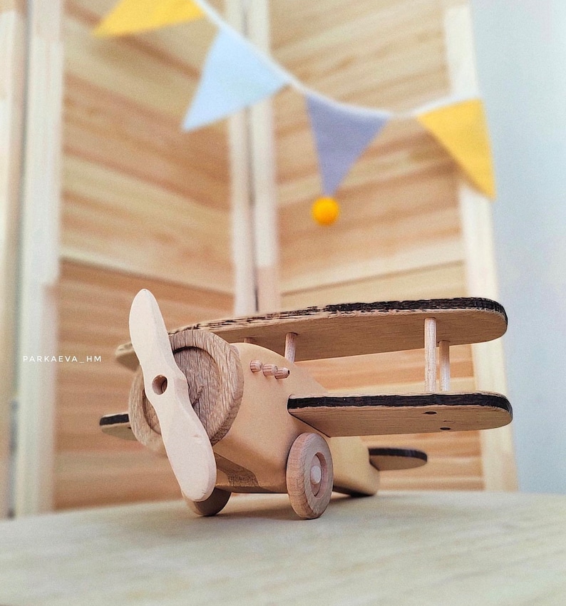 May include: A wooden toy airplane with a propeller, black accents, and wheels. The airplane is sitting on a wooden surface.