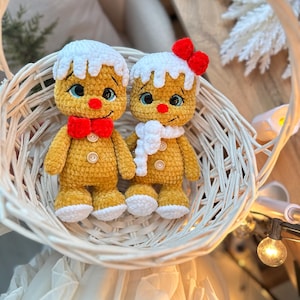 May include: Two crocheted gingerbread man dolls in a white wicker basket. The dolls have yellow bodies, white frosting-like hair, and red bow ties. One doll has a white scarf and a red bow. The dolls have button eyes and are a festive holiday decoration.