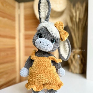 May include: A gray crocheted donkey toy wearing a yellow crocheted dress and a yellow bow. The donkey has blue eyes and a white crocheted mane.