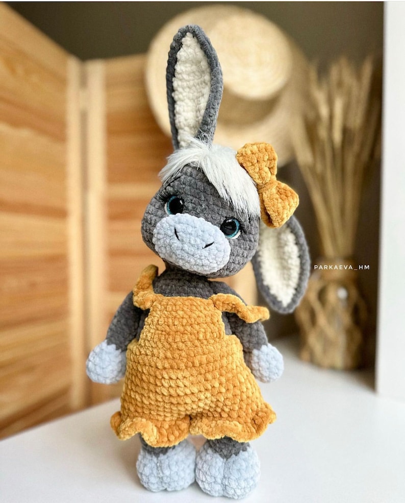 May include: A gray crocheted donkey toy wearing a yellow crocheted dress and a yellow bow. The donkey has blue eyes and a white crocheted mane.