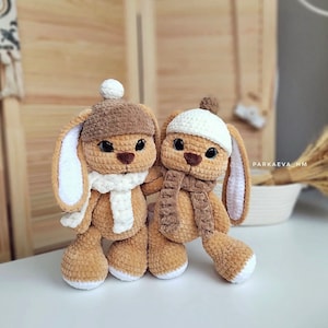 May include: Two crocheted bunny plushies wearing white and brown hats and scarves. The bunnies are brown and white and have black eyes.
