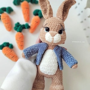 Puede incluir: Un conejito de crochet marr&oacute;n que lleva una chaqueta azul. El conejito tiene grandes ojos negros y una nariz blanca. El conejito est&aacute; de pie sobre una superficie blanca.