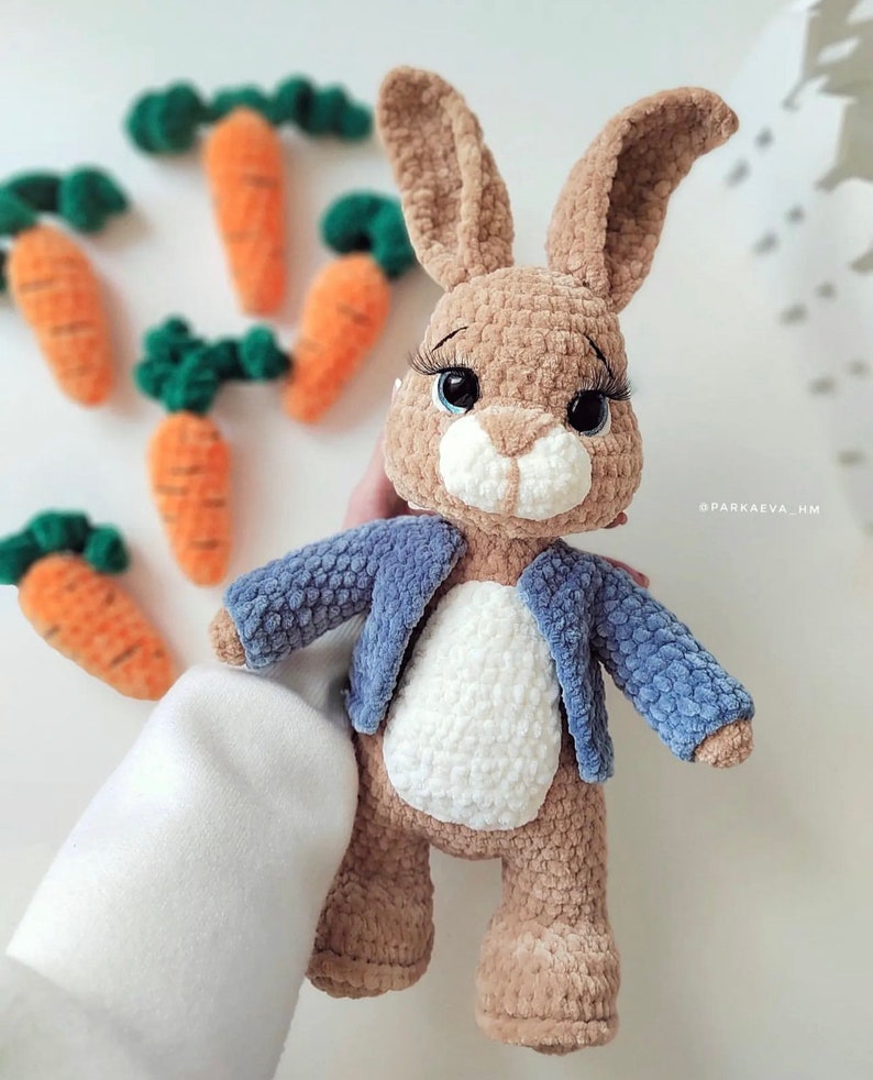 Puede incluir: Un conejito de crochet marr&oacute;n que lleva una chaqueta azul. El conejito tiene grandes ojos negros y una nariz blanca. El conejito est&aacute; de pie sobre una superficie blanca.