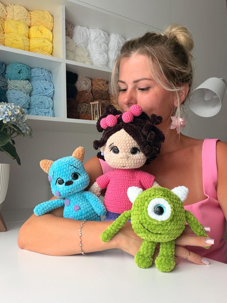 May include: Three handmade crochet dolls: a blue monster with purple spots and orange horns, a girl doll with dark brown curly hair and a pink shirt, and a round green one-eyed monster with white horns, all crafted from soft plush yarn.