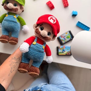 May include: A handmade crochet plush doll of a character wearing a red cap with a white M emblem, a red shirt, blue overalls with yellow buttons, white gloves, and brown shoes. A second green-capped crochet doll is visible in the background.