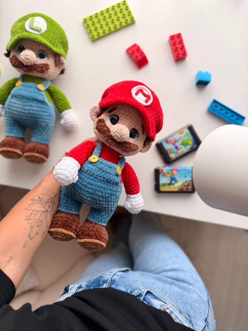 May include: A handmade crochet plush doll of a character wearing a red cap with a white M emblem, a red shirt, blue overalls with yellow buttons, white gloves, and brown shoes. A second green-capped crochet doll is visible in the background.