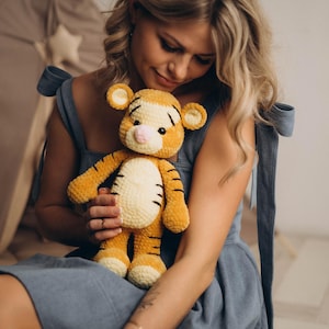 May include: A plush tiger toy crafted from soft, golden-yellow yarn with a cream-coloured belly and muzzle. The stuffed animal features dark brown stripes on its head, back, and limbs, small rounded ears, and a small pink nose with black eyes.