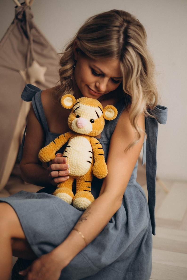 May include: A plush tiger toy crafted from soft, golden-yellow yarn with a cream-coloured belly and muzzle. The stuffed animal features dark brown stripes on its head, back, and limbs, small rounded ears, and a small pink nose with black eyes.