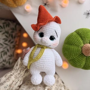Könnte beinhalten: Eine weiß gehäkelte Geisterpuppe mit einem spitzen orangefarbenen Hut, einem hellgrünen Schal und großen dunklen Augen mit blauen Iris. Die Puppe hat einen runden Kopf, eine kleine Nase und rosa Wangen, gefertigt mit sichtbarer Maschenstruktur.