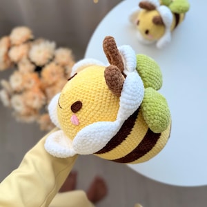 K&ouml;nnte beinhalten: Zwei handgefertigte H&auml;kelbienen. Die Bienen sind gelb mit braunen Streifen und Akzenten, wei&szlig;en und gr&uuml;nen Fl&uuml;geln und braunen Antennen. Eine Biene befindet sich im Vordergrund und eine weitere im Hintergrund. Die Bienen sind weich und pl&uuml;schig und scheinen Spielzeug zu sein.
