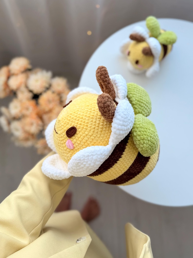 K&ouml;nnte beinhalten: Zwei handgefertigte H&auml;kelbienen. Die Bienen sind gelb mit braunen Streifen und Akzenten, wei&szlig;en und gr&uuml;nen Fl&uuml;geln und braunen Antennen. Eine Biene befindet sich im Vordergrund und eine weitere im Hintergrund. Die Bienen sind weich und pl&uuml;schig und scheinen Spielzeug zu sein.