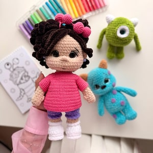 May include: A handmade crochet doll with dark brown curly hair, a pink shirt, purple shorts, and white shoes. Beside it are two smaller crochet monster figures, one green with a single eye and one blue with purple spots, plus a set of colored markers.