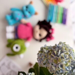 May include: A collection of three crocheted dolls including a blue monster with purple spots, a doll with dark hair and a pink shirt, and a green one-eyed monster, accompanied by a set of colorful markers and a bouquet of blue and green hydrangea flowers.