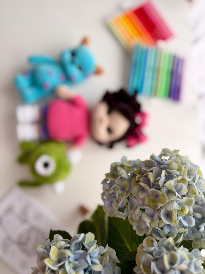 May include: A collection of three crocheted dolls including a blue monster with purple spots, a doll with dark hair and a pink shirt, and a green one-eyed monster, accompanied by a set of colorful markers and a bouquet of blue and green hydrangea flowers.