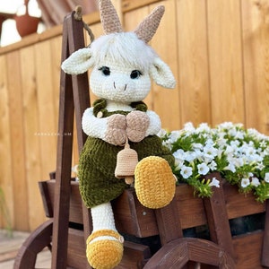 Könnte beinhalten: Eine weiße gehäkelte Ziegenpuppe, die einen grünen Overall mit einer braunen Glocke trägt. Die Puppe sitzt auf einem Holzkarren mit weißen Blumen im Hintergrund.