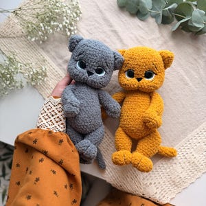 May include: Two plush cat toys made of soft, textured yarn. One cat is dark gray and the other is mustard yellow. Both feature large, round, dark eyes with light blue irises, small black noses, and stitched mouths. The cats have rounded ears.