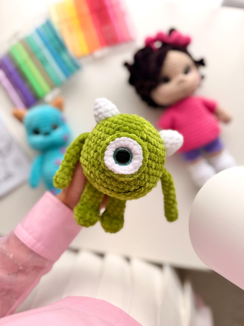 May include: A round, green, crocheted monster toy with a single large central eye, a white iris, and two small white horns on top. The creature has thin arms and legs, a textured yarn surface, and a small stitched mouth.