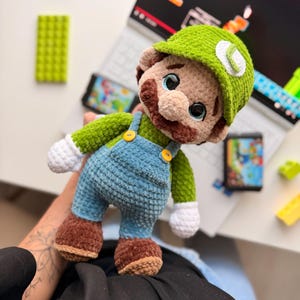 May include: A crocheted plush doll of a character wearing a green cap with a white L emblem, a green long-sleeved shirt, blue overalls with yellow buttons, white gloves, and brown shoes. The doll has a brown mustache and large dark eyes.