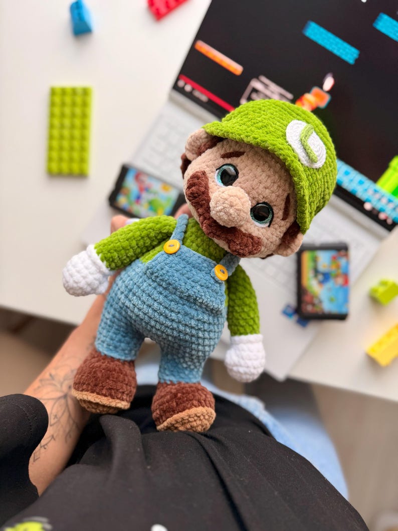 May include: A crocheted plush doll of a character wearing a green cap with a white L emblem, a green long-sleeved shirt, blue overalls with yellow buttons, white gloves, and brown shoes. The doll has a brown mustache and large dark eyes.