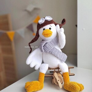 May include: A white crocheted bird wearing a brown aviator hat with goggles and a gray scarf. The bird is sitting on a wooden toy airplane.