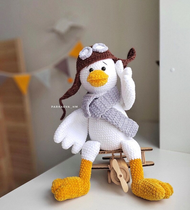 May include: A white crocheted bird wearing a brown aviator hat with goggles and a gray scarf. The bird is sitting on a wooden toy airplane.