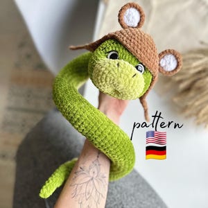 Pode incluir: Brinquedo de cobra de crochê verde com um chapéu de crochê marrom com orelhas brancas. O chapéu tem um pompom branco no topo. A cobra está enrolada e o chapéu está inclinado para o lado. O brinquedo está sendo segurado pela mão de uma pessoa. O texto "pattern" é visível na imagem.
