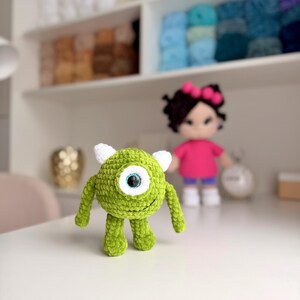 May include: A small, round, green crocheted monster plush toy with a single large central eye, two small white horns on top, and two thin arms and legs. The creature has a textured yarn surface and a stitched smile on its round body.